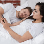 vista-lateral-mulher-verificando-seu-telefone-ao-lado-do-namorado-dormindo