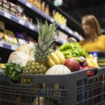 Close up view of shopping cart overloaded with food while in background female person choosing products.