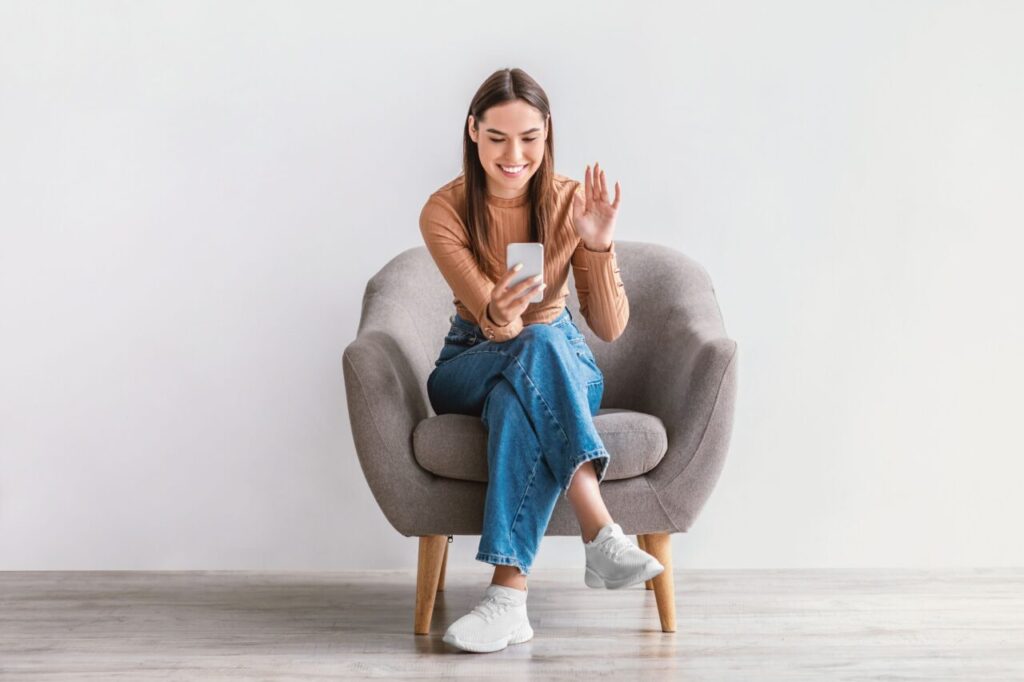 Beautiful,Young,Lady,Sitting,In,Armchair,,Waving,At,Smartphone,Camera,