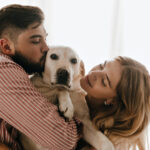 Romantic couple in good mood plays and hugs white dog. Man kisses his labrador against window.