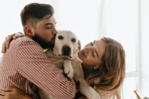 Romantic couple in good mood plays and hugs white dog. Man kisses his labrador against window.