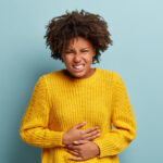 Displeased dark skinned woman suffers from stomachache, period cramps, keeps hands on belly, wears yellow knitted sweater, smirks face, stands against blue background. People and sickness concept