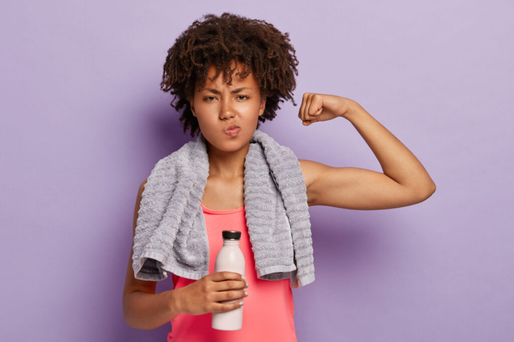 Workout and sport concept. Healthy athletic woman raises arm, shows biceps, wears casual pink top, holds bottle of water, has towel on neck for wiping sweat, exercises indoor, isolated on purple wall