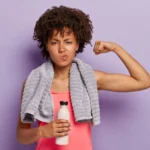 Workout and sport concept. Healthy athletic woman raises arm, shows biceps, wears casual pink top, holds bottle of water, has towel on neck for wiping sweat, exercises indoor, isolated on purple wall