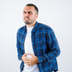 young man having bellyache in checked shirt and white t-shirt and looking exhausted , front view.