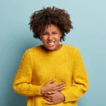 Displeased dark skinned woman suffers from stomachache, period cramps, keeps hands on belly, wears yellow knitted sweater, smirks face, stands against blue background. People and sickness concept