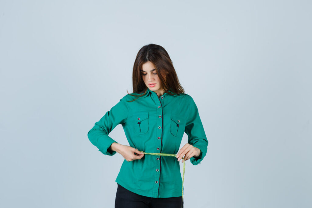 young lady holding measuring tape around her waist in shirt, pants and looking satisfied , front view.