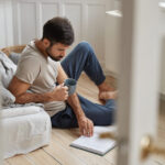 Photo of handsome guy relaxes with literature, enjoy relax at home, concentrated on reading, dressed in casual clothes, sits on floor, holds cup with coffee or tea. People and lifestyle concept