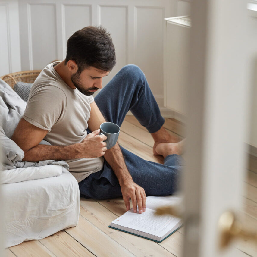 Photo of handsome guy relaxes with literature, enjoy relax at home, concentrated on reading, dressed in casual clothes, sits on floor, holds cup with coffee or tea. People and lifestyle concept