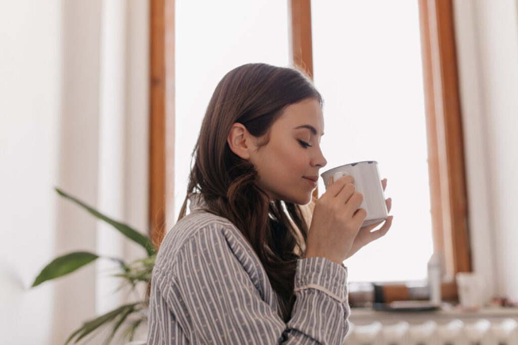 Young woman in striped shirt drinks coffee from iron cup against window. Brunette girl smiling and enjoying tea in cozy apartment