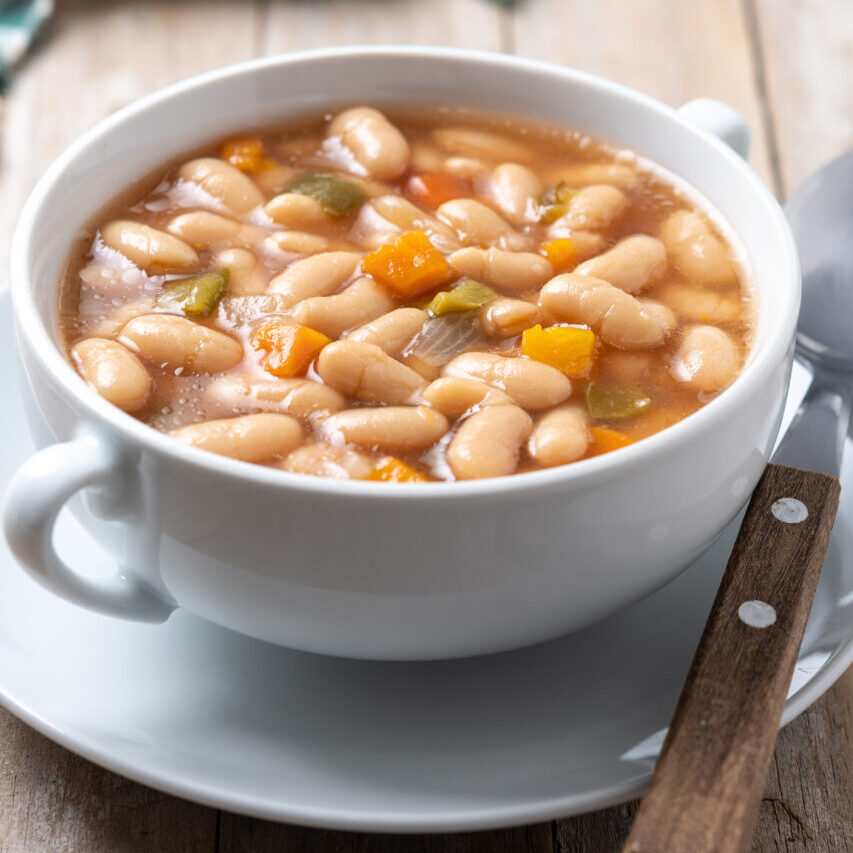 White beans soup with vegetables in white bowl on wooden table.