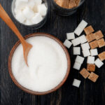 top view of different types and forms of sugar in a bowl and glasses on black wooden background