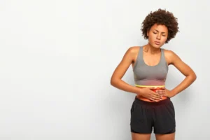 Horizontal shot of dissatisfied African American woman touches belly, suffers from painful cramps, frowns face, dressed in sportclothes, models over white background, free space for your promotion
